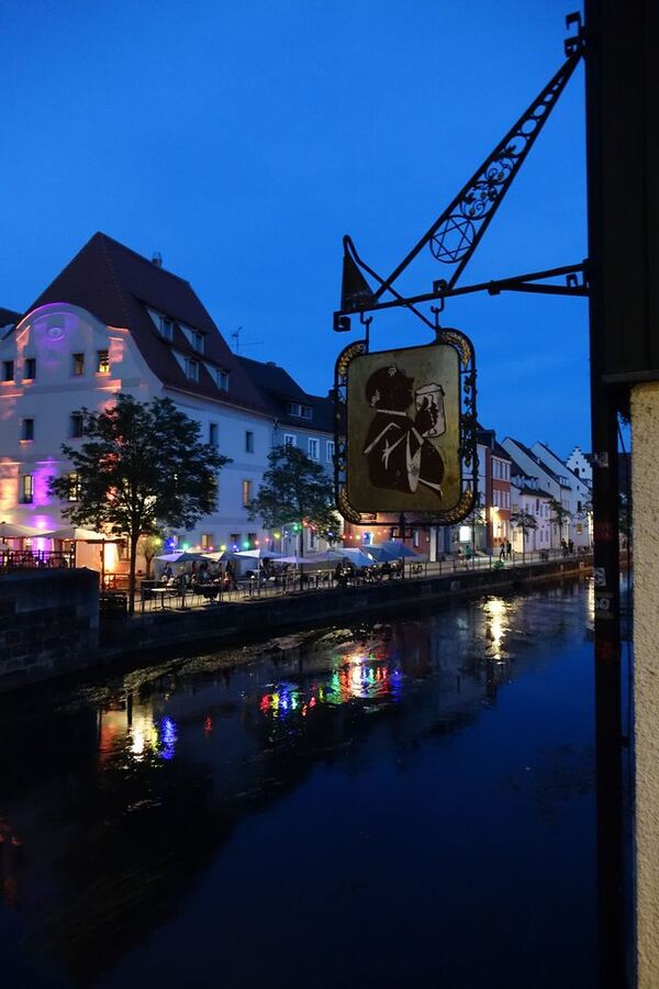 Ein schmiedeeisernes Wirtshausschild ragt über einen Fluss, dahinter beleuchtete Altstadthäuser und Sonnenschirme, die sich im dunklen Wasser spiegeln