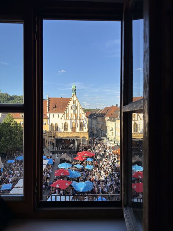 Durch ein geöffnetes Altbaufenster fällt der Blick auf einen Marktplatz mit historischem Rathaus und einem Volksfest mit roten und blauen Sonnenschirmen