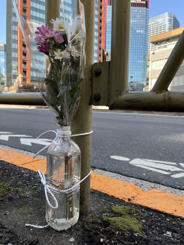 Ein Strauß Chrysanthemen in einer Plastikflasche, festgebunden am Geländer einer Stadtstraße, dahinter Hochhäuser