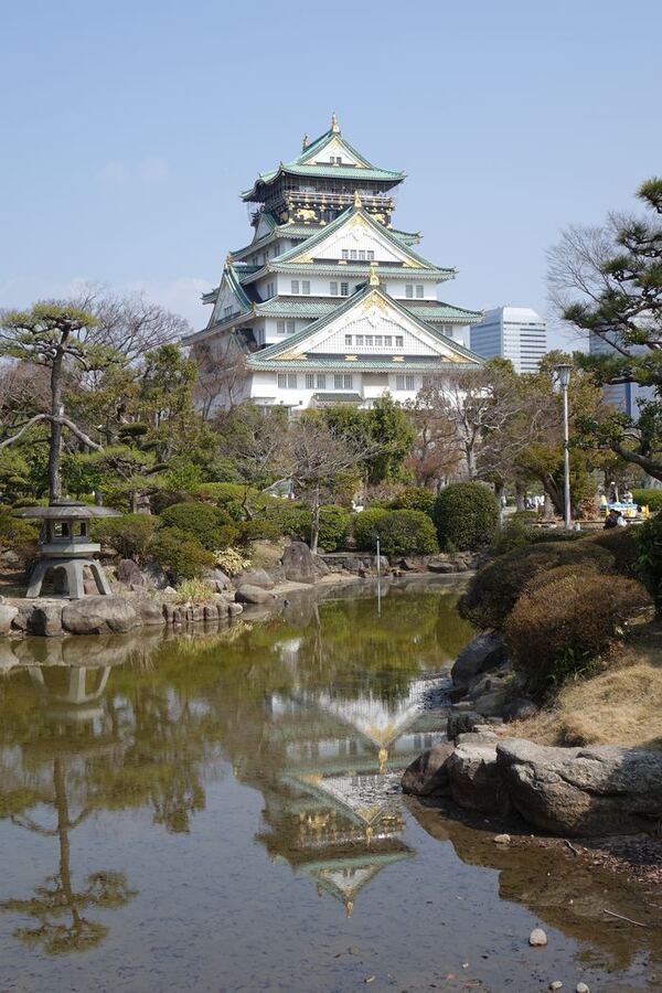 Eine japanische Burg mit geschwungenen Dächern über einem Parkteich, im Vordergrund Kiefern und eine steinerne Laterne