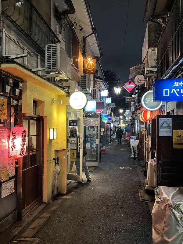Eine schmale Gasse bei Nacht, links und rechts Leuchtreklamen und kleine Lokale, in der Ferne eine einzelne Person