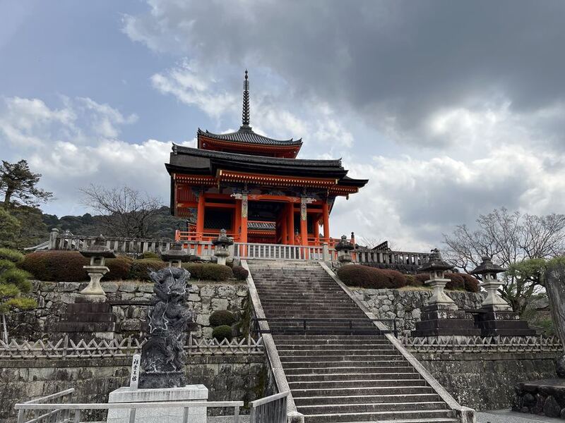 Eine breite Steintreppe führt zu einem rot lackierten Tempeltor mit Pagode, darüber ein bewölkter Himmel