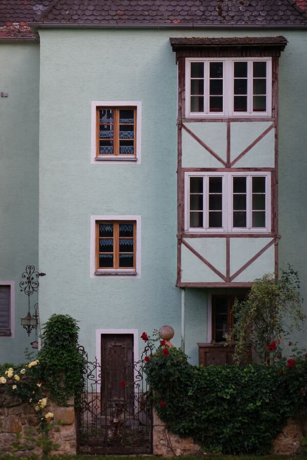 Mintgrüne Hausfassade mit einem Fachwerkerker, davor eine Bruchsteinmauer mit schmiedeeisernem Tor, Efeu und roten und gelben Rosen