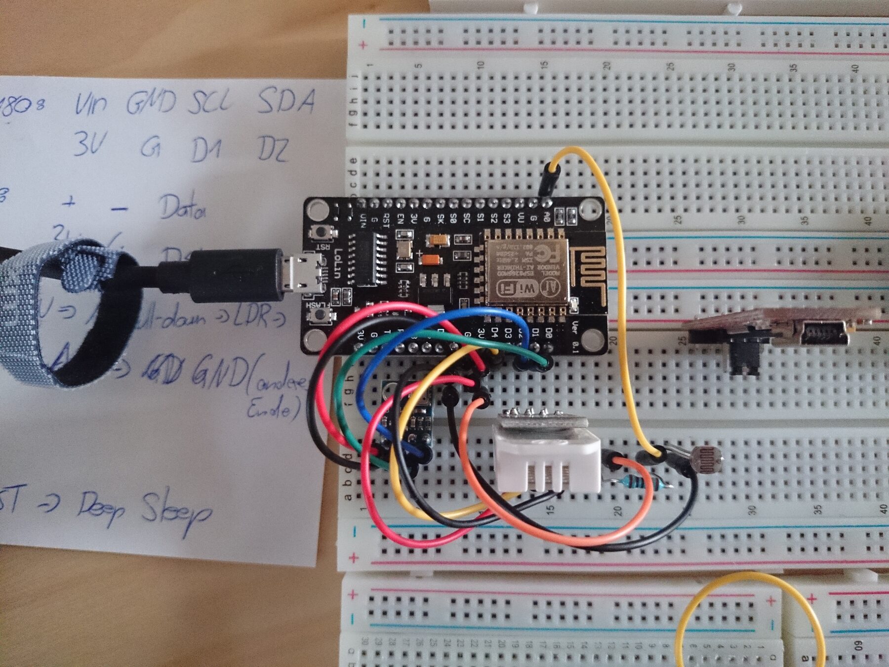 Der Versuchsaufbau auf einem Steckbrett: ein ESP8266-Board am USB-Kabel, bunte Steckbrücken zu den Sensoren und einem Fotowiderstand, links ein Zettel mit handschriftlichen Notizen zur Verkabelung